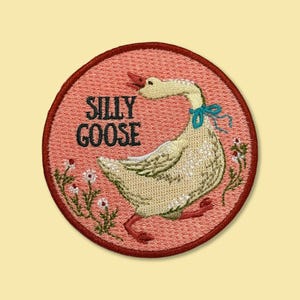 May include: A circular embroidered patch with a coral pink background featuring a cream-coloured goose wearing a blue ribbon around its neck. The text SILLY GOOSE is stitched in black letters to the left, accompanied by small white flowers.