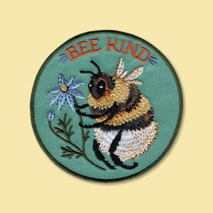 May include: A circular embroidered patch featuring a light blue background with a central bumblebee illustration and a blue flower. The text BEE KIND is stitched in orange at the top. The bee has black, yellow, and white thread details.