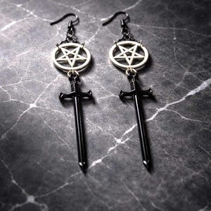 May include: A pair of black earrings with a gothic theme. Each earring features a silver pentagram pendant, a black sword charm, and a black ear hook. The earrings are displayed on a dark, marbled surface.