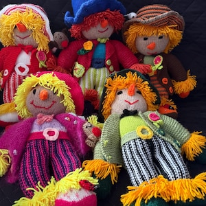 May include: A group of handmade knitted dolls with yarn hair and fabric clothing. The dolls have smiling faces and are dressed in various outfits, including jackets, hats, and striped trousers. The colour palette includes red, pink, green, and yellow.
