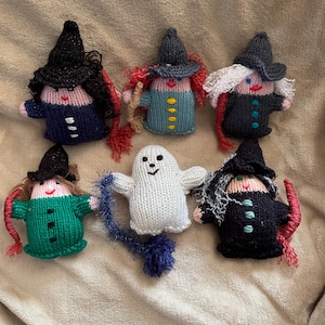 May include: A collection of six handmade Halloween-themed knitted dolls. The dolls include five witches with pointed hats and a white ghost. The witches wear various colored outfits, and the ghost has a smiling face and a blue tassel.