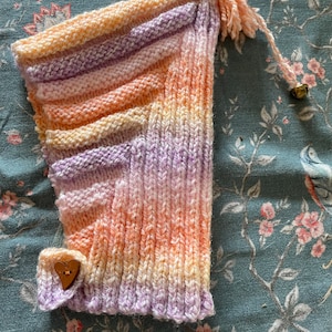 May include: A hand-knitted pixie hat in pastel shades of peach, lavender, and cream, with a wooden heart-shaped button and a small bell. The hat has a pointed top and a decorative tassel. The knitted design features a gradient of colours.