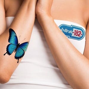 May include: A blue butterfly tattoo on an arm. The butterfly has black detailing and is positioned on the inner arm. A white tube top is visible. The text "blink-182" is on a blue and white patch.