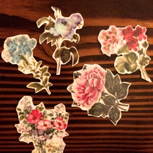 K&ouml;nnte beinhalten: F&uuml;nf Papierblumen-Verzierungen mit floralen Designs. Die Blumen sind in den Farben Rosa, Blau, Rot und Wei&szlig; gehalten. Die Blumen befinden sich auf einem braunen Holzgrund.