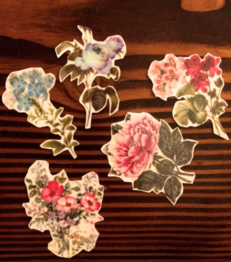 K&ouml;nnte beinhalten: F&uuml;nf Papierblumen-Verzierungen mit floralen Designs. Die Blumen sind in den Farben Rosa, Blau, Rot und Wei&szlig; gehalten. Die Blumen befinden sich auf einem braunen Holzgrund.