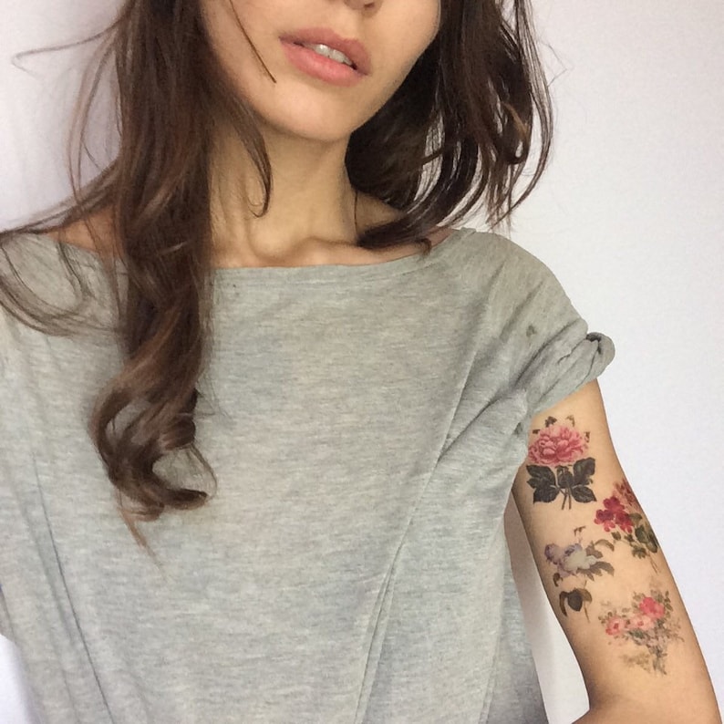 K&ouml;nnte beinhalten: Eine Frau mit langen braunen Haaren tr&auml;gt ein graues T-Shirt mit floralen tempor&auml;ren Tattoos auf ihrem Oberarm.