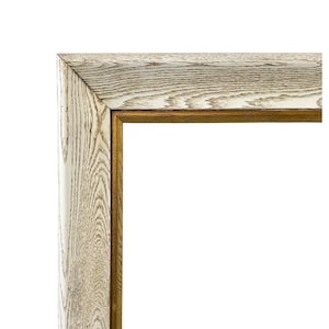 Puede incluir: Primer plano de una esquina de un marco de fotos de madera. El marco tiene un acabado claro, blanquecino, con veta de madera visible. Un borde interior fino de color dorado bordea la abertura, creando un contraste con la madera más clara.