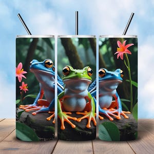 May include: Three stainless steel tumblers with a vibrant frog design. The frogs are blue and green with orange feet, perched on a log with pink flowers and a forest background. Each tumbler has a black straw.
