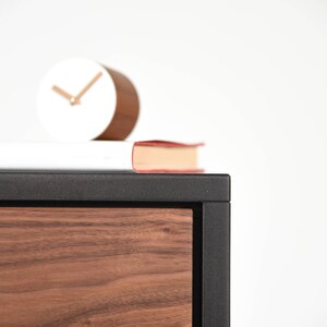May include: A black and wood cabinet with a drawer. A white clock and a red book are on top of the cabinet.