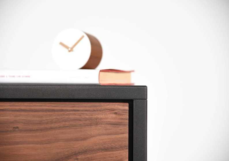 May include: A black and wood cabinet with a drawer. A white clock and a red book are on top of the cabinet.