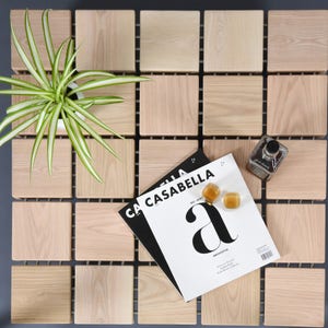 May include: A square wooden table with a grid pattern, featuring a potted plant, magazines, and decorative items. The table is made of light-colored wood blocks, arranged in a grid. The magazines are titled "CASABELLA".