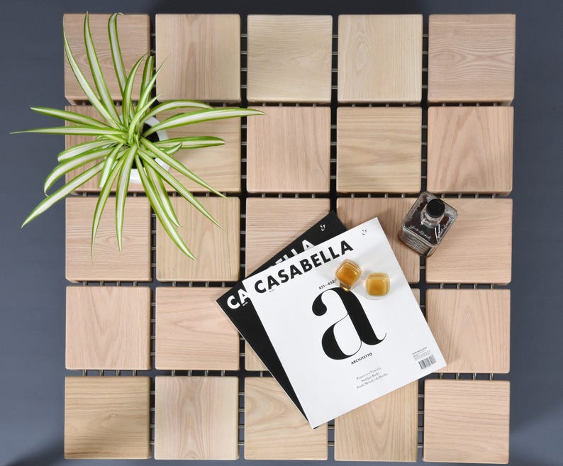 May include: A square wooden table with a grid pattern, featuring a potted plant, magazines, and decorative items. The table is made of light-colored wood blocks, arranged in a grid. The magazines are titled "CASABELLA".