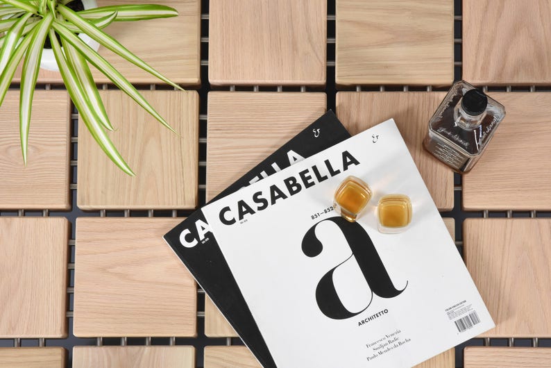 May include: A still-life composition featuring a magazine titled "CASABELLA" with a large "A" on the cover, two small square glass containers, and a glass bottle with a black cap. The items are arranged on a light wood surface with a plant.