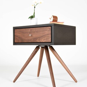 May include: A dark brown nightstand with a single drawer and a wood grain finish. The nightstand has four legs that are angled outward. A white orchid plant is sitting on top of the nightstand.
