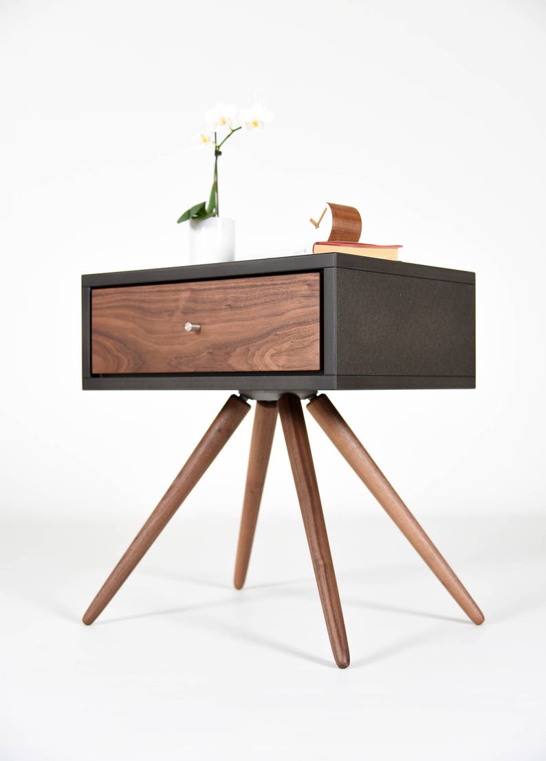 May include: A dark brown nightstand with a single drawer and a wood grain finish. The nightstand has four legs that are angled outward. A white orchid plant is sitting on top of the nightstand.