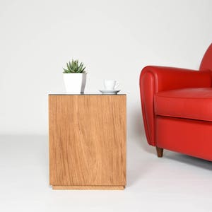 May include: A wooden side table with a glass top. A small potted plant and a white cup and saucer are on the table. A red armchair is in the background.