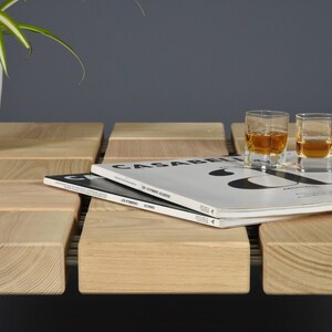 May include: A wooden coffee table with a grid-like design, featuring two shot glasses of amber liquid, a bottle of whiskey, and a stack of magazines. A white potted plant is on the left. The table is made of light-colored wood.