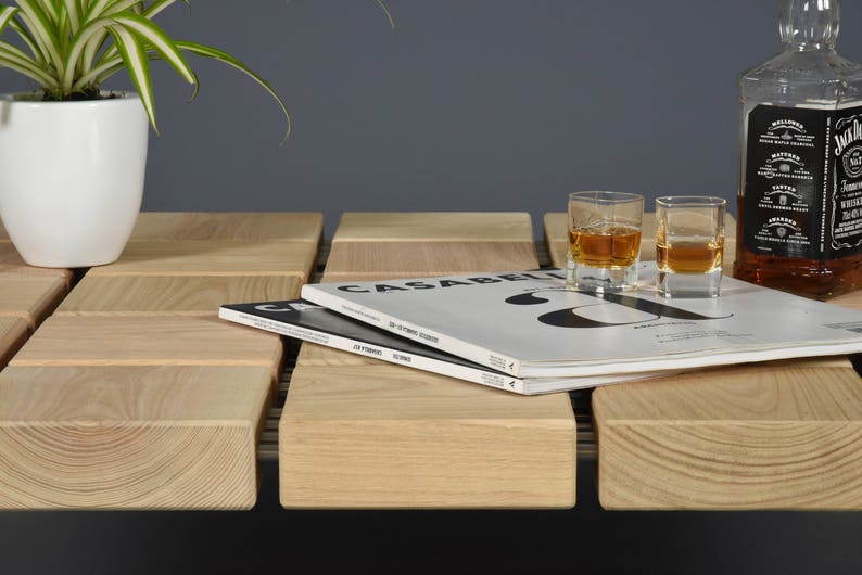 May include: A wooden coffee table with a grid-like design, featuring two shot glasses of amber liquid, a bottle of whiskey, and a stack of magazines. A white potted plant is on the left. The table is made of light-colored wood.