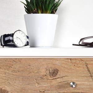 May include: A white dresser with a wooden drawer. The drawer has a light brown wood grain pattern and a silver knob. A silver watch with a black and white striped band is on top of the dresser. A small potted plant with green leaves is also on top of the dresser. A pair of black glasses is on the dresser.