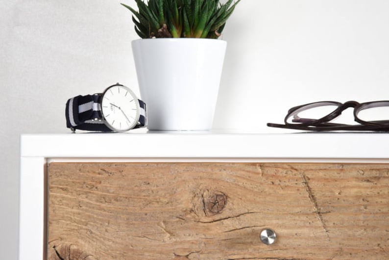May include: A white dresser with a wooden drawer. The drawer has a light brown wood grain pattern and a silver knob. A silver watch with a black and white striped band is on top of the dresser. A small potted plant with green leaves is also on top of the dresser. A pair of black glasses is on the dresser.