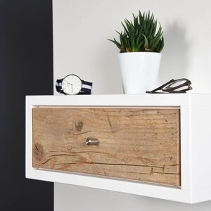 May include: White floating bedside table with a single drawer. The drawer front is made of reclaimed wood with a natural finish. A watch and a pair of glasses are on the top surface. A small potted succulent plant is on the right side of the shelf.