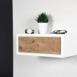 May include: White floating bedside table with a single drawer. The drawer front is made of wood with a natural finish. A watch and a pair of glasses are on top of the bedside table.