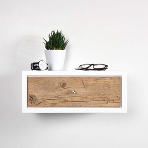 May include: White floating shelf with a single drawer. The drawer front is made of wood with a natural wood grain finish. A small potted plant, a watch, and a pair of glasses are on top of the shelf.