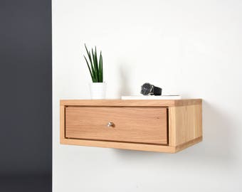 May include: A light wood floating nightstand with a single drawer and a silver knob. The nightstand is mounted to a white wall and has a small potted plant and a watch on top.