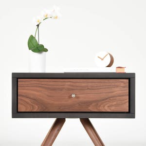 May include: A dark wood nightstand with a single drawer and black trim. The drawer has a silver knob. The nightstand has angled legs and is sitting on a white surface.