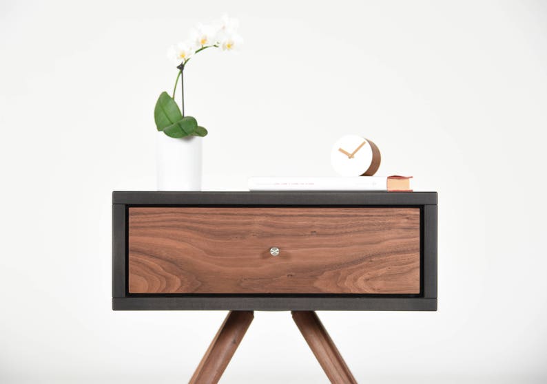 May include: A dark wood nightstand with a single drawer and black trim. The drawer has a silver knob. The nightstand has angled legs and is sitting on a white surface.