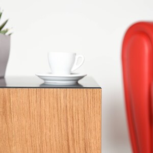 May include: A wooden end table with a glass top. A white coffee cup and saucer are on the table. A red armchair is in the background.
