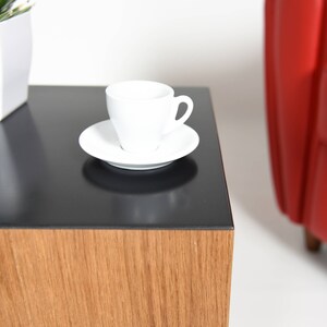 May include: A black and wood end table with a white cup and saucer on top. The table has a black top and a wood base.
