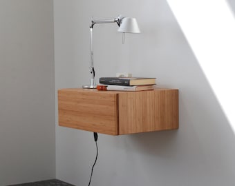 May include: A light wood floating nightstand with a drawer and a lamp on top. The nightstand is mounted to a white wall.