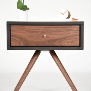 May include: A dark wood nightstand with a single drawer and three angled legs. The drawer is made of light wood with a wood grain pattern. The nightstand is on a white background.