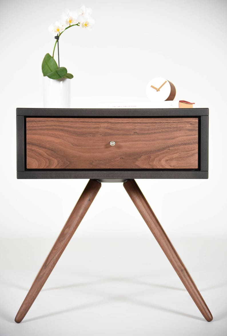 May include: A dark wood nightstand with a single drawer and three angled legs. The drawer is made of light wood with a wood grain pattern. The nightstand is on a white background.