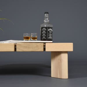 May include: A light-colored wooden coffee table with a rectangular top and a single leg. A bottle of whiskey, two glasses of whiskey, a plant in a white pot, and a magazine are on the table.