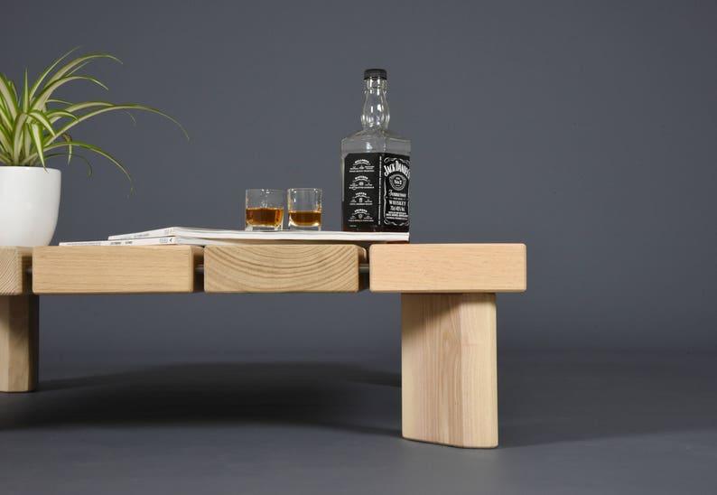 May include: A light-colored wooden coffee table with a rectangular top and a single leg. A bottle of whiskey, two glasses of whiskey, a plant in a white pot, and a magazine are on the table.