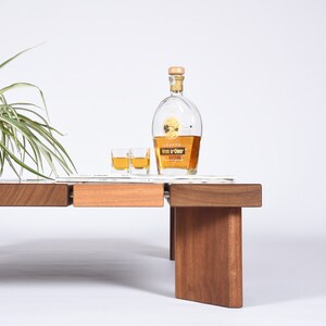May include: A wooden coffee table with a light brown finish and a glass top. Two small glasses of liquor and a bottle of Grappa Vite d'Oro liqueur are on the table.