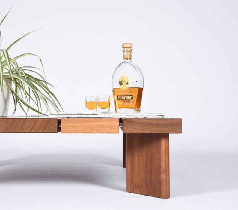May include: A wooden coffee table with a light brown finish and a glass top. Two small glasses of liquor and a bottle of Grappa Vite d'Oro liqueur are on the table.