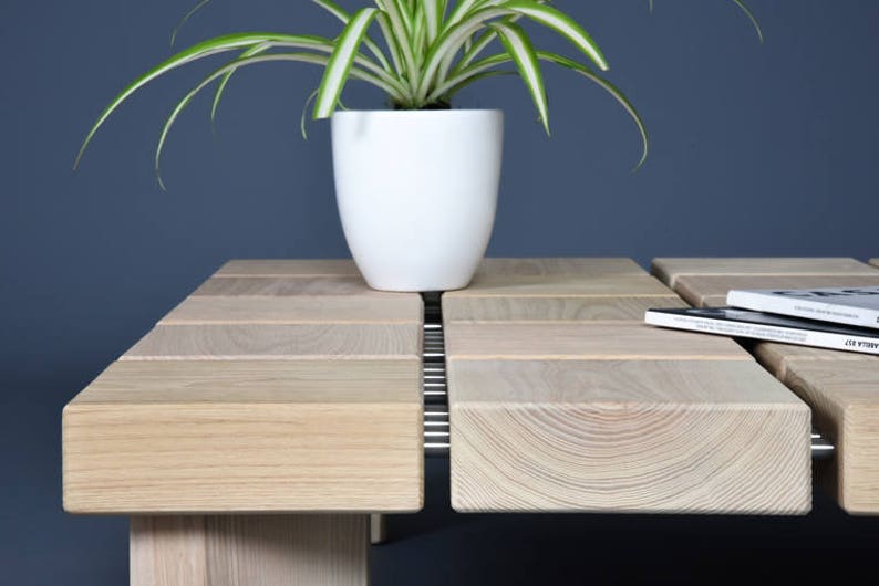 May include: A light-colored wooden coffee table with a modern design. The table features a series of rectangular wooden blocks, with a white ceramic pot holding a green plant. A stack of books rests on the table.