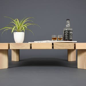 May include: A light-colored wooden coffee table with a rectangular design. The table has a white potted plant, a bottle of whiskey, two glasses, and a small stack of papers on top. The table is set against a gray background.