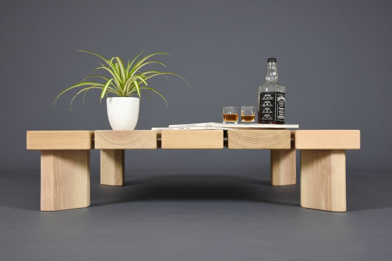 May include: A light-colored wooden coffee table with a rectangular design. The table has a white potted plant, a bottle of whiskey, two glasses, and a small stack of papers on top. The table is set against a gray background.