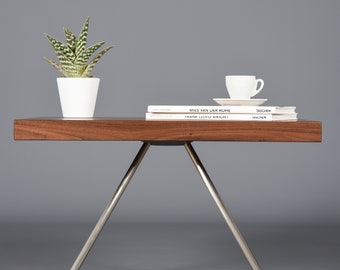 Può includere: Un tavolo in legno con finitura in noce e tre gambe argentate. Il tavolo ha una piccola pianta grassa in vaso e una pila di libri sopra. I libri sono intitolati "Mies van der Rohe" e "Frank Lloyd Wright".
