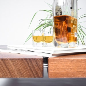 May include: A wooden table with a stack of magazines on top. A bottle of amber liquid and two shot glasses filled with the same liquid are on the magazines. A green plant is in the background.