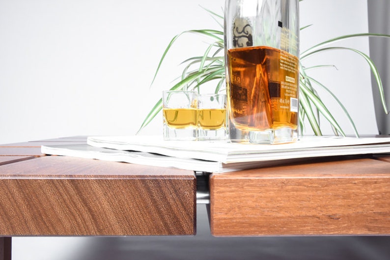 May include: A wooden table with a stack of magazines on top. A bottle of amber liquid and two shot glasses filled with the same liquid are on the magazines. A green plant is in the background.