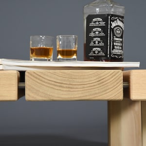 May include: A still life arrangement featuring a bottle of Jack Daniel's whiskey, two shot glasses filled with amber liquid, and a stack of magazines. The scene is set on a light-colored wooden surface with a gray backdrop.
