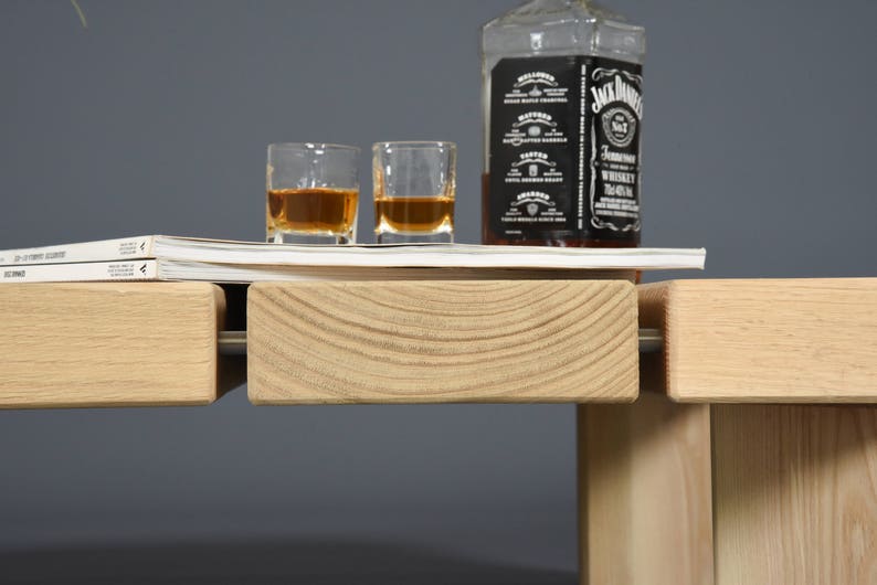 May include: A still life arrangement featuring a bottle of Jack Daniel's whiskey, two shot glasses filled with amber liquid, and a stack of magazines. The scene is set on a light-colored wooden surface with a gray backdrop.