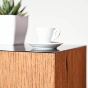 May include: A wooden end table with a black glass top. A white cup and saucer are on the table, and a potted plant is in the background.