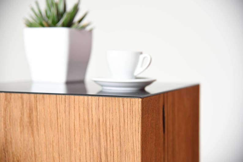 May include: A wooden end table with a black glass top. A white cup and saucer are on the table, and a potted plant is in the background.