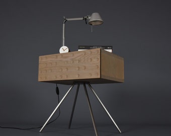 Puede incluir: Una mesita de noche de madera con una tapa texturizada única y tres patas de metal. La mesa tiene un pequeño reloj y un libro en la parte superior.